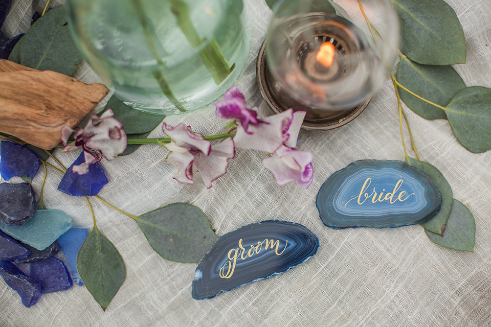 San diego beach wedding at blacks beach white blanket set up on the sand with rocks behind and light blue decorative pillows with light blue and pink flower decor and crystal wine glasses with blue geode rock name plates for bride and groom in calligraphy writing wedding photo idea for beach blanket set up