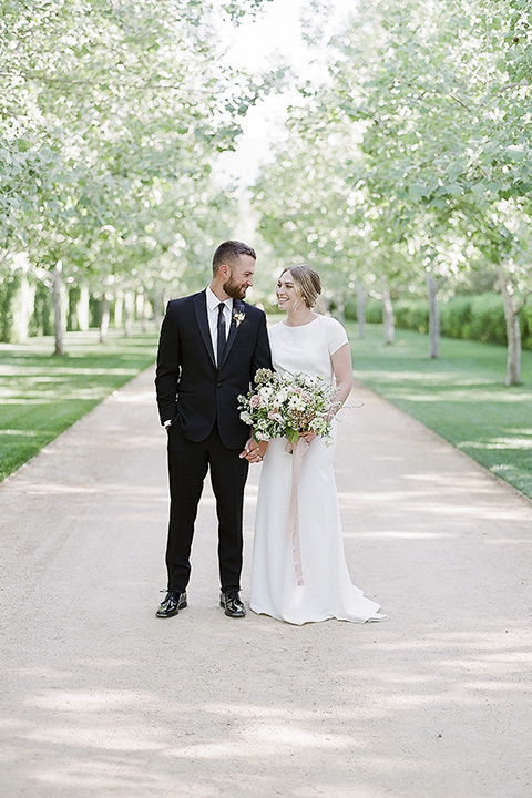 Santa barbara outdoor black tie wedding at kestrel park bride simple form fitting gown with a high neckline and short sleeves and groom black peak lapel tuxedo and a white dress shirt with a long black skinny tie and green floral boutonniere holding hands and bride holding white and green floral bridal bouquet