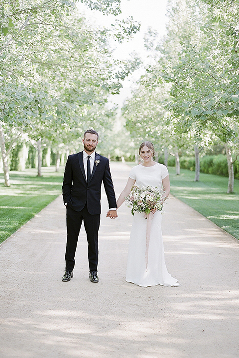 Santa barbara outdoor black tie wedding at kestrel park bride simple form fitting gown with a high neckline and short sleeves and groom black peak lapel tuxedo and a white dress shirt with a long black skinny tie and green floral boutonniere holding hands and bride holding white and green floral bridal bouquet