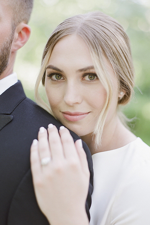 Santa barbara outdoor black tie wedding at kestrel park bride simple form fitting gown with a high neckline and short sleeves and groom black peak lapel tuxedo and a white dress shirt with a long black skinny tie and green floral boutonniere hugging bride behind groom close up on bride