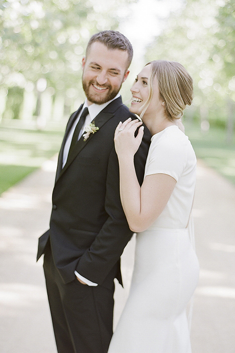 Santa barbara outdoor black tie wedding at kestrel park bride simple form fitting gown with a high neckline and short sleeves and groom black peak lapel tuxedo and a white dress shirt with a long black skinny tie and green floral boutonniere hugging bride behind groom smiling