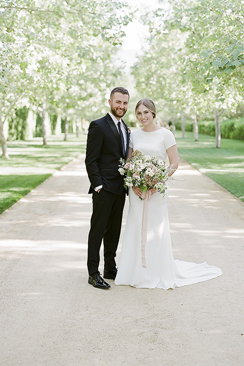 Santa barbara outdoor black tie wedding at kestrel park bride simple form fitting gown with a high neckline and short sleeves and groom black peak lapel tuxedo and a white dress shirt with a long black skinny tie and green floral boutonniere hugging and smiling bride holding white and green floral bridal bouquet