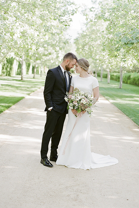 Santa barbara outdoor black tie wedding at kestrel park bride simple form fitting gown with a high neckline and short sleeves and groom black peak lapel tuxedo and a white dress shirt with a long black skinny tie and green floral boutonniere hugging and touching heads bride holding white and green floral bridal bouquet