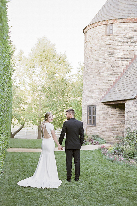 Santa barbara outdoor black tie wedding at kestrel park bride simple form fitting gown with a high neckline and short sleeves and groom black peak lapel tuxedo and a white dress shirt with a long black skinny tie and green floral boutonniere holding hands far away back view