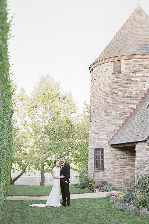 Santa barbara outdoor black tie wedding at kestrel park bride simple form fitting gown with a high neckline and short sleeves and groom black peak lapel tuxedo and a white dress shirt with a long black skinny tie and green floral boutonniere hugging far away