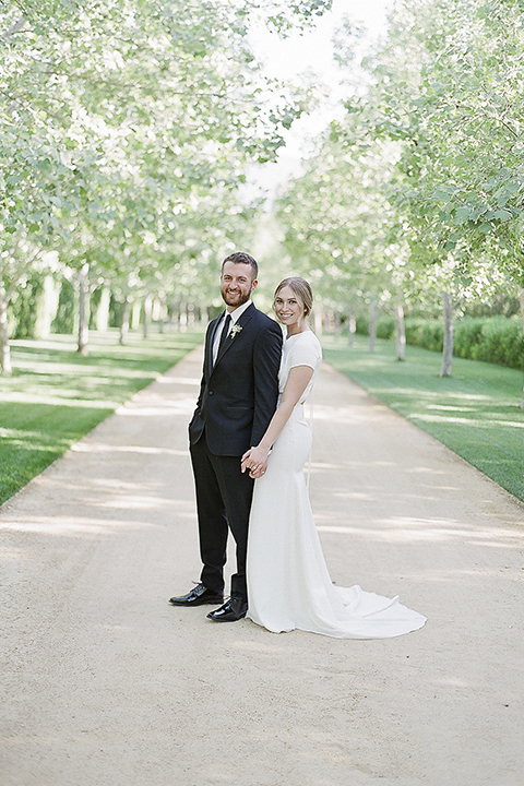 Santa barbara outdoor black tie wedding at kestrel park bride simple form fitting gown with a high neckline and short sleeves and groom black peak lapel tuxedo and a white dress shirt with a long black skinny tie and green floral boutonniere holding hands bride behind groom smiling