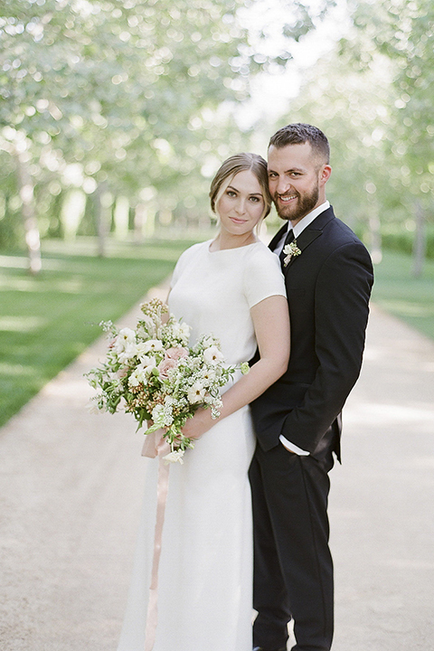 Santa barbara outdoor black tie wedding at kestrel park bride simple form fitting gown with a high neckline and short sleeves and groom black peak lapel tuxedo and a white dress shirt with a long black skinny tie and green floral boutonniere hugging and bride holding white and green floral bridal bouquet