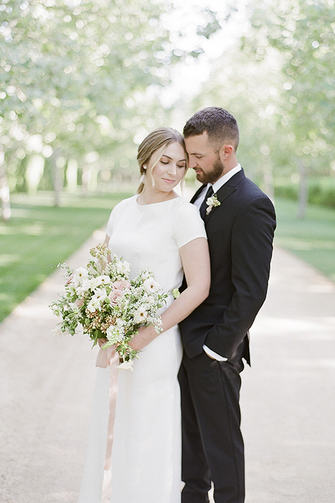 Santa barbara outdoor black tie wedding at kestrel park bride simple form fitting gown with a high neckline and short sleeves and groom black peak lapel tuxedo and a white dress shirt with a long black skinny tie and green floral boutonniere hugging and touching heads bride holding white and green floral bridal bouquet