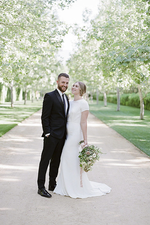 Santa barbara outdoor black tie wedding at kestrel park bride simple form fitting gown with a high neckline and short sleeves and groom black peak lapel tuxedo and a white dress shirt with a long black skinny tie and green floral boutonniere hugging and smiling bride holding white and green floral bridal bouquet