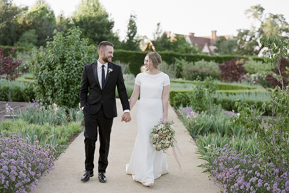 Santa barbara outdoor black tie wedding at kestrel park bride simple form fitting gown with a high neckline and short sleeves and groom black peak lapel tuxedo and a white dress shirt with a long black skinny tie and green floral boutonniere walking and holding hands bride holding white and green floral bridal bouquet