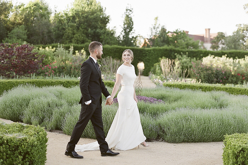 Santa barbara outdoor black tie wedding at kestrel park bride simple form fitting gown with a high neckline and short sleeves and groom black peak lapel tuxedo and a white dress shirt with a long black skinny tie and green floral boutonniere holding hands and walking and bride holding white and green floral bridal bouquet