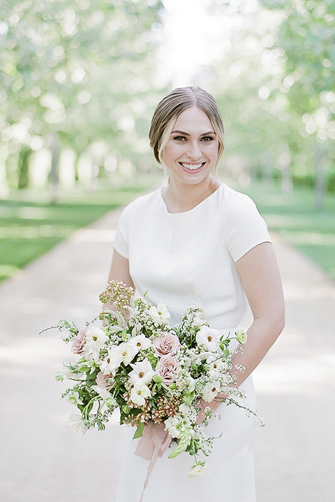 Santa barbara outdoor black tie wedding at kestrel park bride simple form fitting gown with a high neckline and short sleeves holding white and green floral bridal bouquet smiling