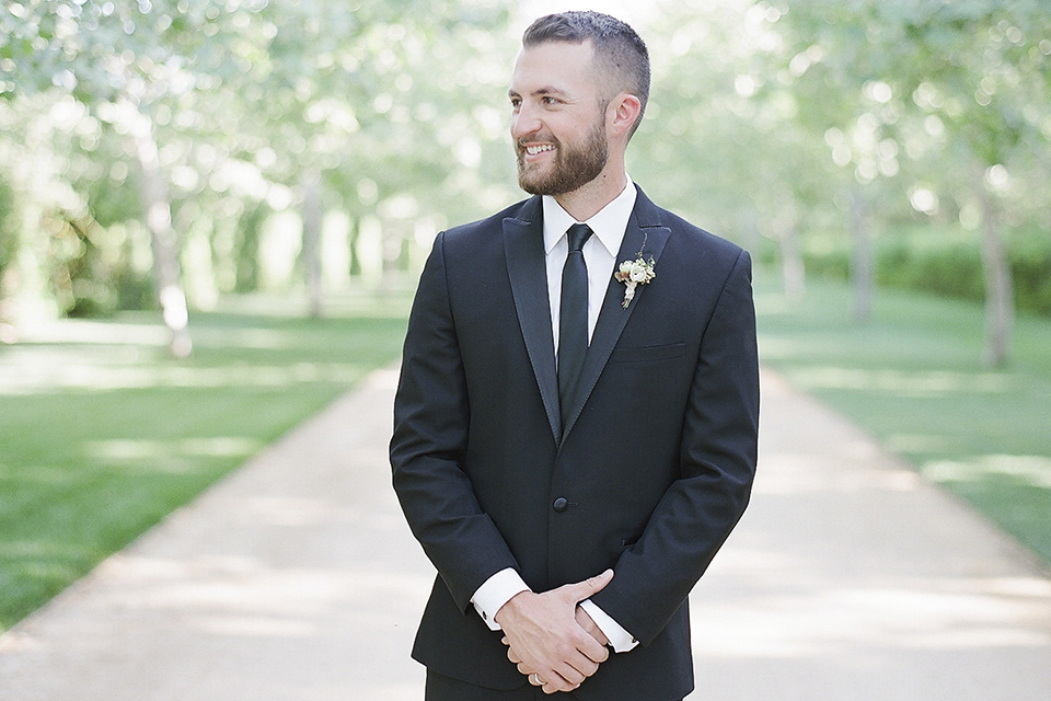 Santa barbara outdoor black tie wedding at kestrel park groom black peak lapel tuxedo with a white dress shirt and long black skinny tie with a green floral boutonniere standing with hands crossed in front smiling close up