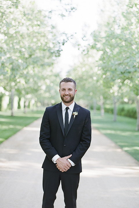 Santa barbara outdoor black tie wedding at kestrel park groom black peak lapel tuxedo with a white dress shirt and long black skinny tie with a green floral boutonniere standing with hands crossed in front smiling