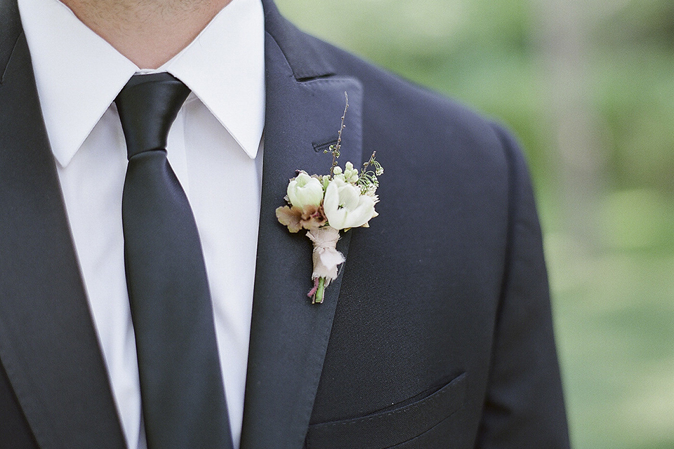 Santa barbara outdoor black tie wedding at kestrel park groom black peak lapel tuxedo with a white dress shirt and long black skinny tie with a green floral boutonniere close up