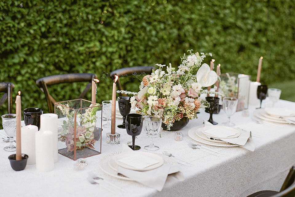 Santa barbara outdoor black tie wedding at kestrel park table set up with white lace linen and white place settings with white linen napkins and black wine glasses with white and green floral centerpiece decor with black and gold candles and wooden rustic chairs