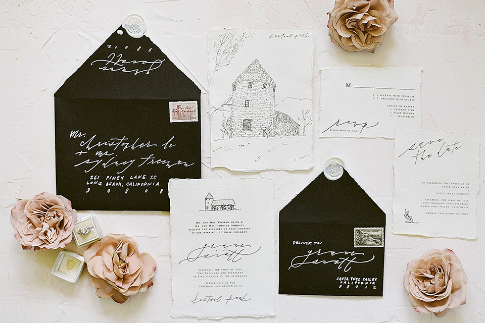 Santa barbara outdoor black tie wedding at kestrel park white background with black and white wedding invitations with black envelopes and white invitations with black calligraphy writing and drawing of venue with light pink flower decor on ground next to invitations wedding photo idea for invitations