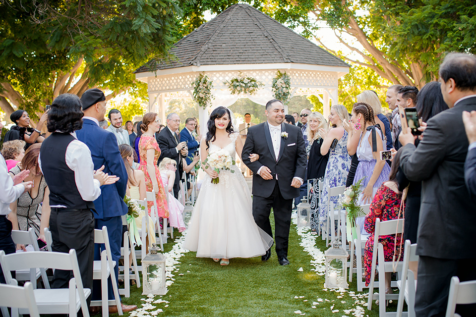 Orange county outdoor summer wedding at the heritage museum bride ball gown with lace straps and a sweetheart neckline with a short tulle skirt and long veil with groom black shawl lapel tuxedo with a white dress shirt and white vest with a white tie and white floral boutonniere holding hands and walking down the aisle after ceremony bride holding white floral bridal bouquet