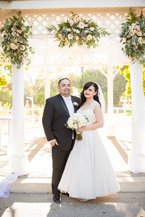 Orange county outdoor summer wedding at the heritage museum bride ball gown with lace straps and a sweetheart neckline with a short tulle skirt and long veil with groom black shawl lapel tuxedo with a white dress shirt and white vest with a white tie and white floral boutonniere standing and hugging bride holding white floral bridal bouquet