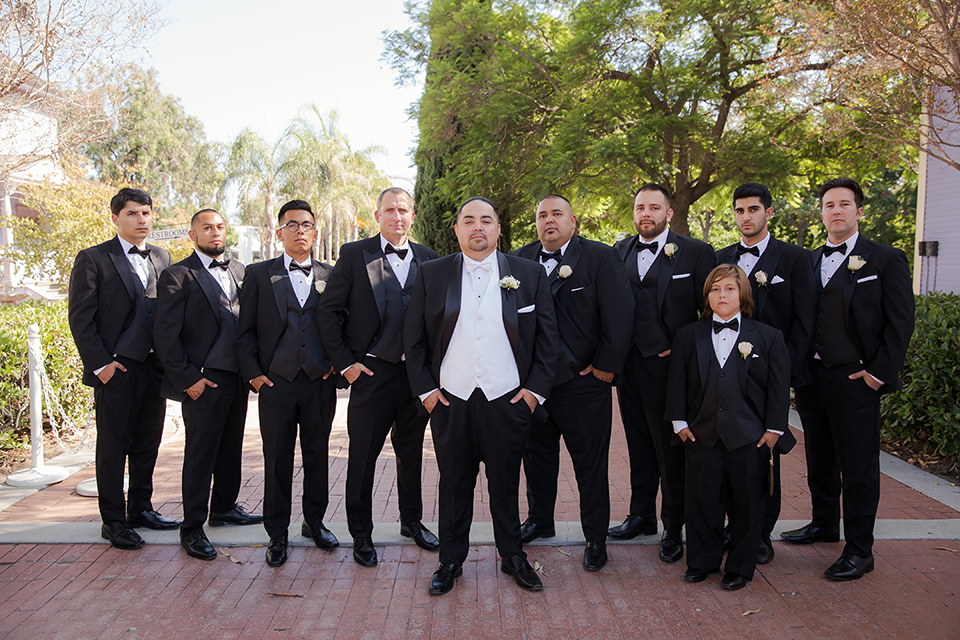 Orange county outdoor summer wedding at the heritage museum groom black shawl lapel tuxedo with a white dress shirt and white vest with a white tie and white floral boutonniere standing with groomsmen and ring bearer black tuxedos with black bow ties