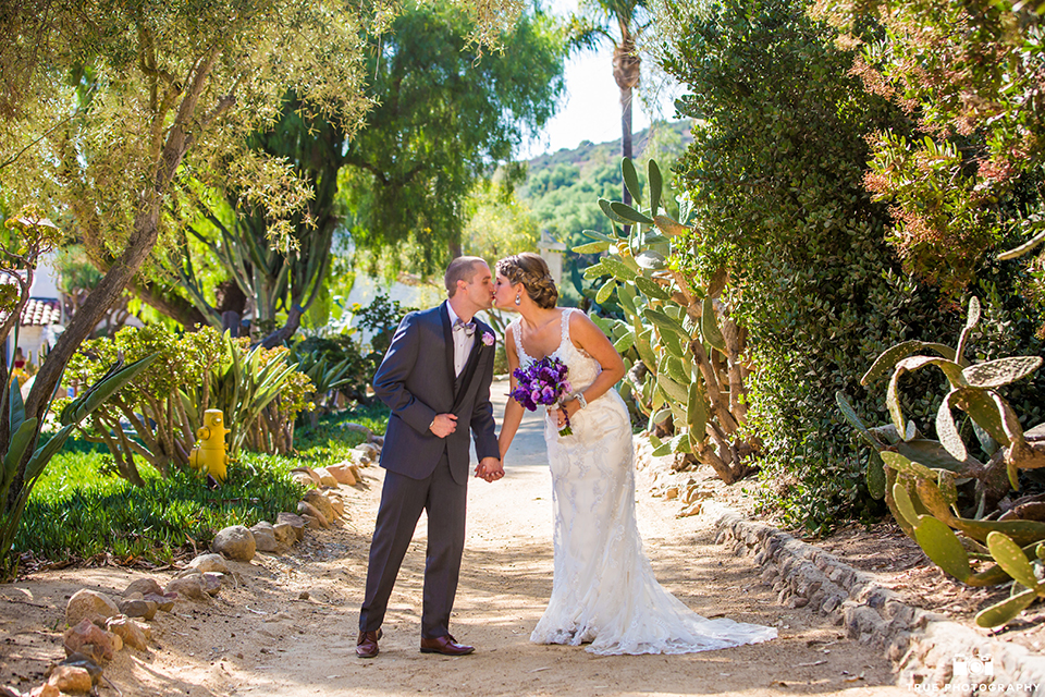 San diego outdoor wedding at leo carillo ranch bride form fitting gown with a sweetheart neckline with thin straps and beaded detail on bodice with groom charcoal grey tuxedo with a black shawl and matching vest with a white dress shirt and plaid bow tie with a white floral boutonniere holding hands and kissing