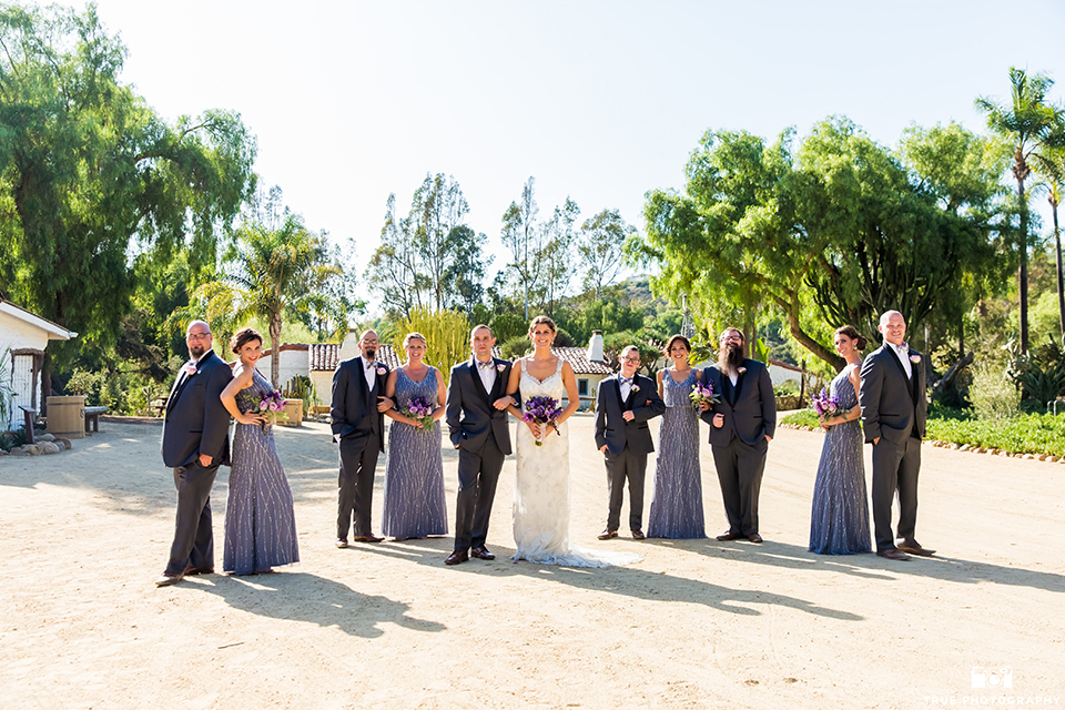 San diego outdoor wedding at leo carillo ranch bride form fitting gown with a sweetheart neckline with thin straps and beaded detail on bodice with groom charcoal grey tuxedo with a black shawl and matching vest with a white dress shirt and plaid bow tie with a white floral boutonniere standing with wedding party groomsmen charcoal grey tuxedos and bridesmaids long dusty blue beaded dresses