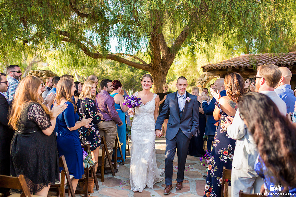 San diego outdoor wedding at leo carillo ranch bride form fitting gown with a sweetheart neckline with thin straps and beaded detail on bodice with groom charcoal grey tuxedo with a black shawl and matching vest with a white dress shirt and plaid bow tie with a white floral boutonniere walking down the aisle after ceremony holding hands and smiling