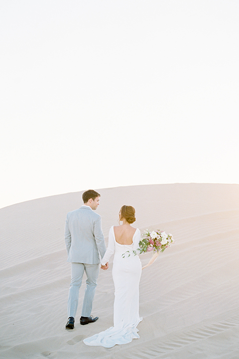 California outdoor wedding shoot at glamis sand dunes bride form fitting long sleeved gown with an open back design and plunging neckline with low hair updo and groom light grey peak lapel suit with a white dress shirt and no tie casual look holding hands and bride holding white and light pink floral bridal bouquet 