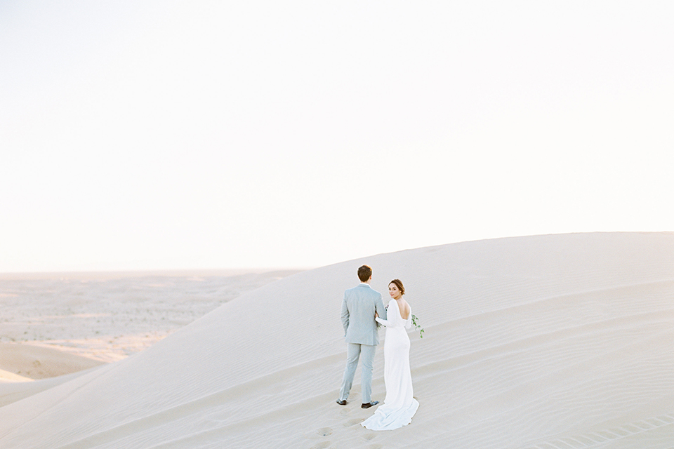 California outdoor wedding shoot at glamis sand dunes bride form fitting long sleeved gown with an open back design and plunging neckline with low hair updo and groom light grey peak lapel suit with a white dress shirt and no tie casual look holding hands far away