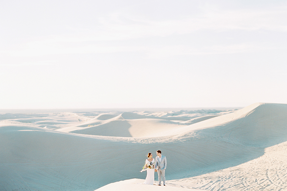 California outdoor wedding shoot at glamis sand dunes bride form fitting long sleeved gown with an open back design and plunging neckline with low hair updo and groom light grey peak lapel suit with a white dress shirt and no tie casual look holding hands and bride holding white and light pink floral bridal bouquet