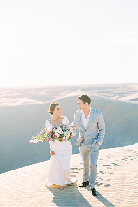 California outdoor wedding shoot at glamis sand dunes bride form fitting long sleeved gown with an open back design and plunging neckline with low hair updo and groom light grey peak lapel suit with a white dress shirt and no tie casual look holding hands and bride holding white and pink floral bridal bouquet
