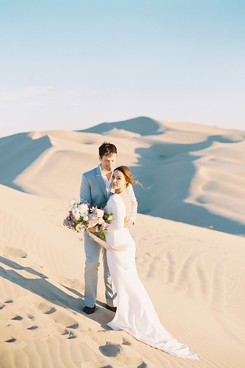 California outdoor wedding shoot at glamis sand dunes bride form fitting long sleeved gown with an open back design and plunging neckline with low hair updo and groom light grey peak lapel suit with a white dress shirt and no tie casual look hugging and bride holding white and pink floral bridal bouquet
