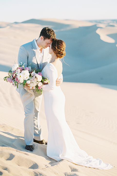 California outdoor wedding shoot at glamis sand dunes bride form fitting long sleeved gown with an open back design and plunging neckline with low hair updo and groom light grey peak lapel suit with a white dress shirt and no tie casual look hugging and bride holding white and light pink floral bridal bouquet