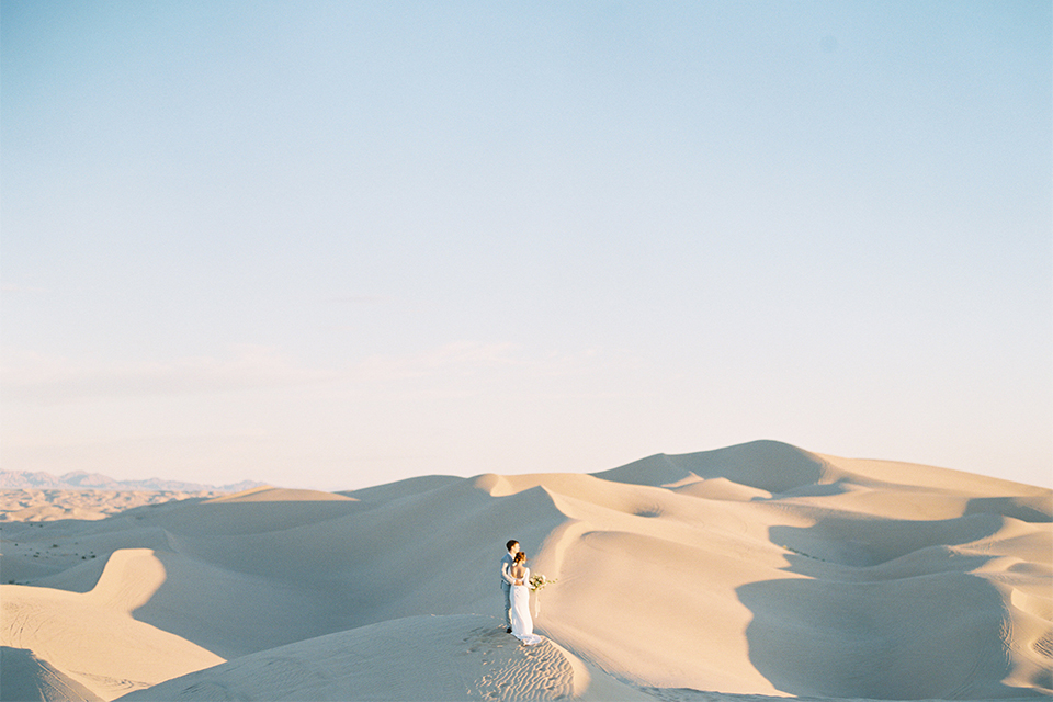 California outdoor wedding shoot at glamis sand dunes bride form fitting long sleeved gown with an open back design and plunging neckline with low hair updo and groom light grey peak lapel suit with a white dress shirt and no tie casual look hugging far away and bride holding white and light pink floral bridal bouquet