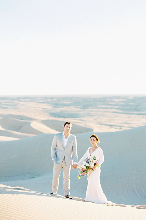 California outdoor wedding shoot at glamis sand dunes bride form fitting long sleeved gown with an open back design and plunging neckline with low hair updo and groom light grey peak lapel suit with a white dress shirt and no tie casual look standing and holding hands and bride holding white and light pink floral bridal bouquet