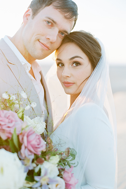 California outdoor wedding shoot at glamis sand dunes bride form fitting long sleeved gown with an open back design and plunging neckline with low hair updo and groom light grey peak lapel suit with a white dress shirt and no tie casual look hugging close up and touching heads bride holding white and light pink floral bridal bouquet