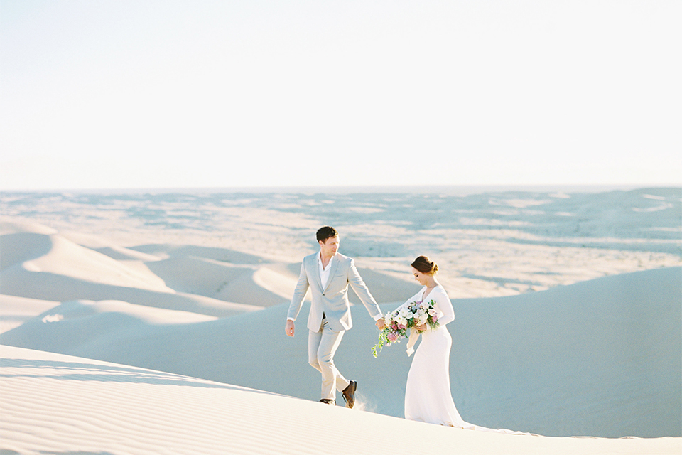 California outdoor wedding shoot at glamis sand dunes bride form fitting long sleeved gown with an open back design and plunging neckline with low hair updo and groom light grey peak lapel suit with a white dress shirt and no tie casual look holding hands and walking bride holding white and light pink floral bridal bouquet 