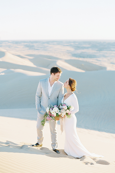 California outdoor wedding shoot at glamis sand dunes bride form fitting long sleeved gown with an open back design and plunging neckline with low hair updo and groom light grey peak lapel suit with a white dress shirt and no tie casual look standing and holding hands and bride holding white and light pink floral bridal bouquet