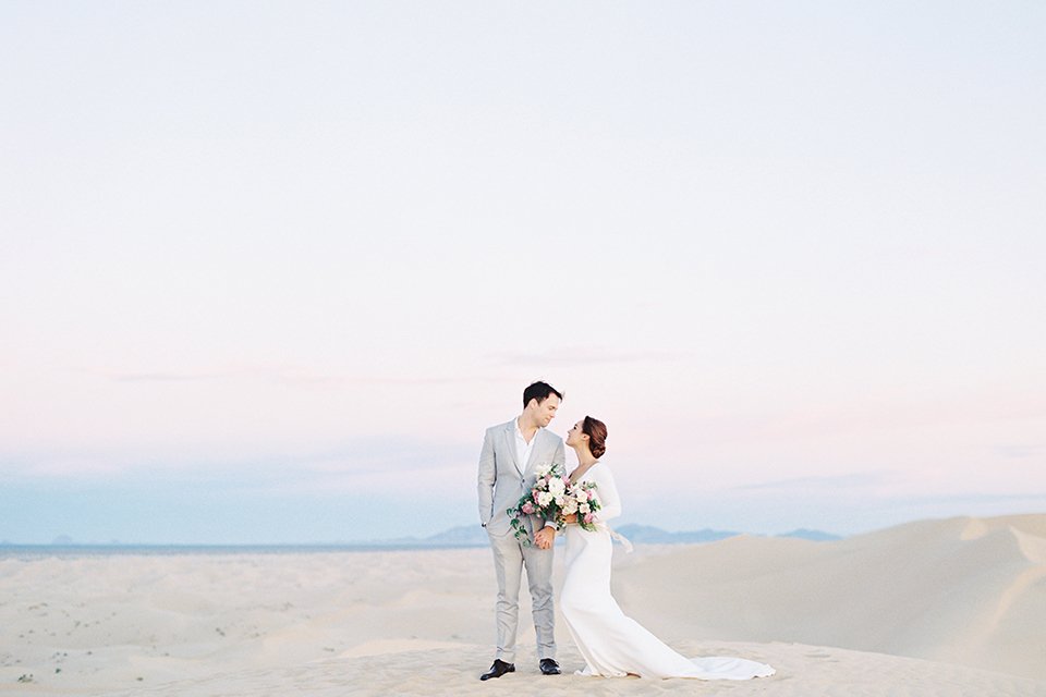California outdoor wedding shoot at glamis sand dunes bride form fitting long sleeved gown with an open back design and plunging neckline with low hair updo and groom light grey peak lapel suit with a white dress shirt and no tie casual look hugging far away and bride holding white and light pink floral bridal bouquet