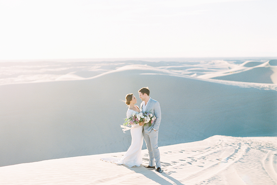 California outdoor wedding shoot at glamis sand dunes bride form fitting long sleeved gown with an open back design and plunging neckline with low hair updo and groom light grey peak lapel suit with a white dress shirt and no tie casual look hugging and bride holding white and light pink floral bridal bouquet