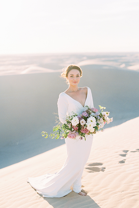 California outdoor wedding shoot at glamis sand dunes bride form fitting long sleeved gown with an open back design and plunging neckline with low hair updo holding white and light pink floral bridal bouquet