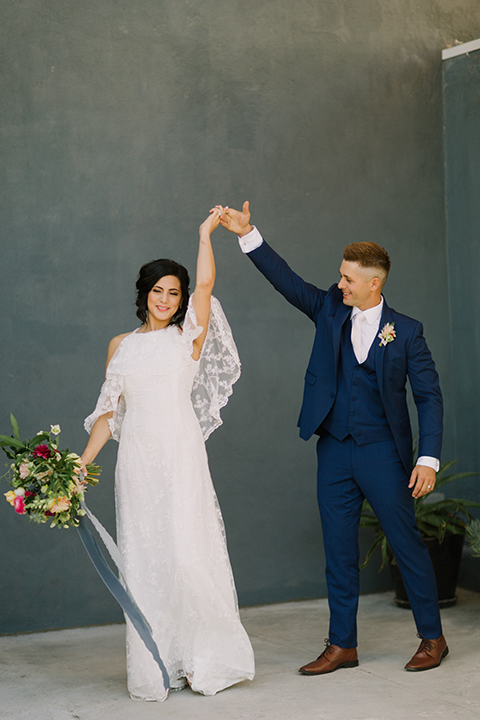 Los angeles outdoor colorful wedding bride a line gown with a high neckline and open shoulder lace sleeves and groom cobalt blue notch lapel suit by allure men with a matching vest and white dress shirt with a long white matte tie and white floral boutonniere dancing and twirling bride