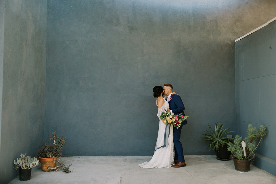 Los angeles outdoor colorful wedding bride a line gown with a high neckline and open shoulder lace sleeves and groom cobalt blue notch lapel suit by allure men with a matching vest and white dress shirt with a long white matte tie and white floral boutonniere first look bride and groom kissing