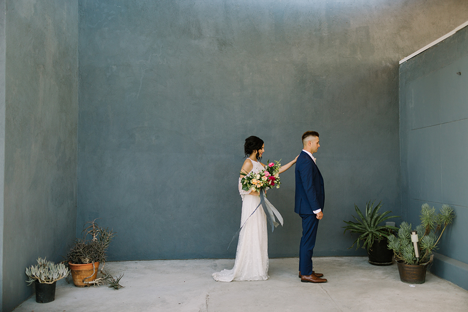 Los angeles outdoor colorful wedding bride a line gown with a high neckline and open shoulder lace sleeves and groom cobalt blue notch lapel suit by allure men with a matching vest and white dress shirt with a long white matte tie and white floral boutonniere first look bride tapping groom on the shoulder