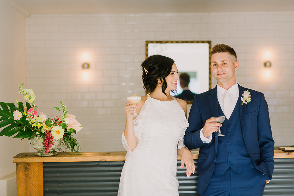 Los angeles outdoor colorful wedding bride a line gown with a high neckline and open shoulder lace sleeves and groom cobalt blue notch lapel suit by allure men with a matching vest and white dress shirt with a long white matte tie and white floral boutonniere standing by the bar holding drinks