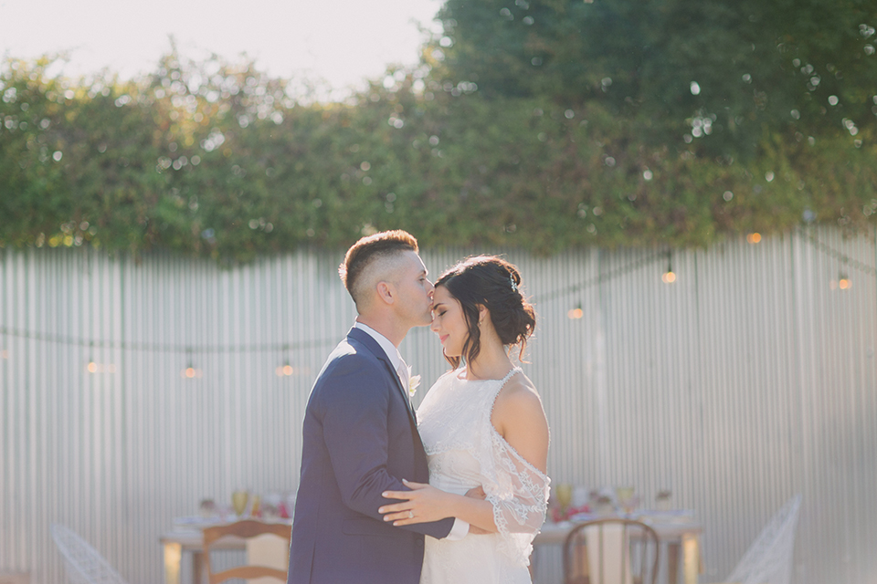 Los angeles outdoor colorful wedding bride a line gown with a high neckline and open shoulder lace sleeves and groom cobalt blue notch lapel suit by allure men with a matching vest and white dress shirt with a long white matte tie and white floral boutonniere standing and hugging groom kissing brides forehead