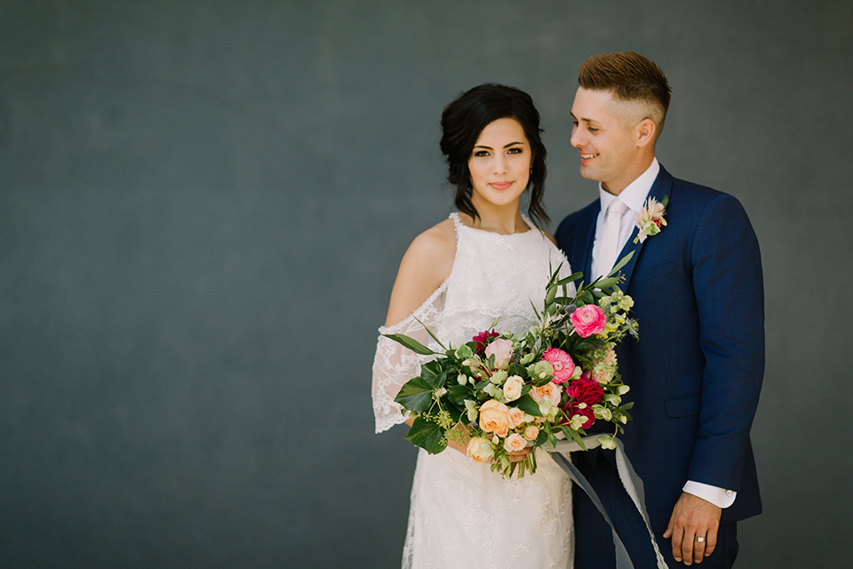 Los angeles outdoor colorful wedding bride a line gown with a high neckline and open shoulder lace sleeves and groom cobalt blue notch lapel suit by allure men with a matching vest and white dress shirt with a long white matte tie and white floral boutonniere standing and hugging bride holding colorful floral bridal bouquet