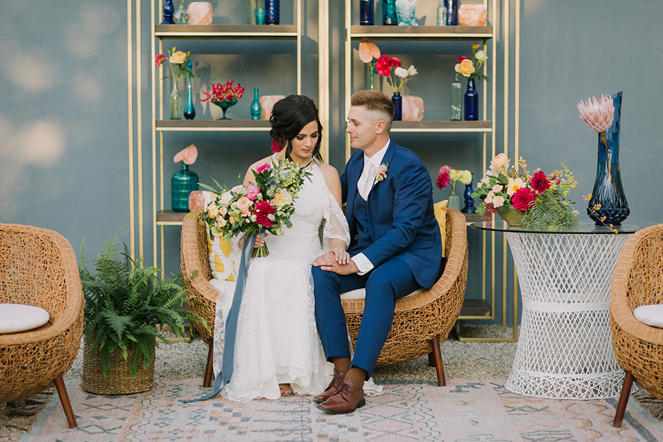 Los angeles outdoor colorful wedding bride a line gown with a high neckline and open shoulder lace sleeves and groom cobalt blue notch lapel suit by allure men with a matching vest and white dress shirt with a long white matte tie and white floral boutonniere sitting on wicker style couch with flower decor in background bride holding colorful floral bouquet