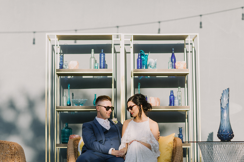 Los angeles outdoor colorful wedding bride a line gown with a high neckline and open shoulder lace sleeves and groom cobalt blue notch lapel suit by allure men with a matching vest and white dress shirt with a long white matte tie and white floral boutonniere sitting on wicker style couch wearing sunglasses