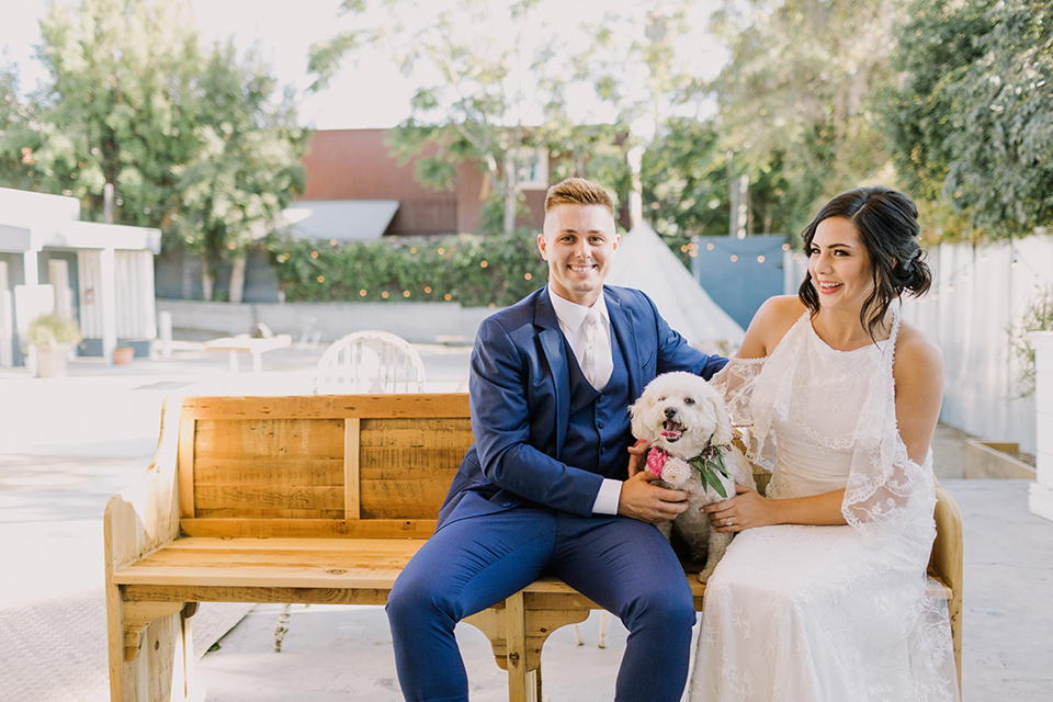 Los angeles outdoor colorful wedding bride a line gown with a high neckline and open shoulder lace sleeves and groom cobalt blue notch lapel suit by allure men with a matching vest and white dress shirt with a long white matte tie and white floral boutonniere sitting on wooden bench with dog smiling